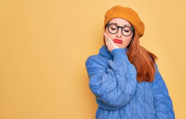 Fransız beresi takan, sarı arka planda gözlük takan genç ve güzel bir kızıl kadın kollarını kavuşturup depresyondan sıkılmış ve yorgun görünüyor..