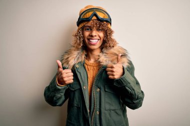 Kıvırcık saçlı, kar sporu kıyafetleri ve kayak gözlüğü takan, başarılı el hareketi yapan, gülümseyen ve mutlu genç Afrikalı Amerikalı kayakçı kadın. Neşeli ifade ve kazanan jest.