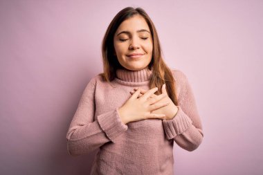 Güzel genç bir kadın pembe izole edilmiş arka planda balıkçı yaka kazak giyiyor. Elleri göğsünde, gözleri kapalı gülümsüyor ve yüzünde minnet ifadesi var. Sağlık kavramı.