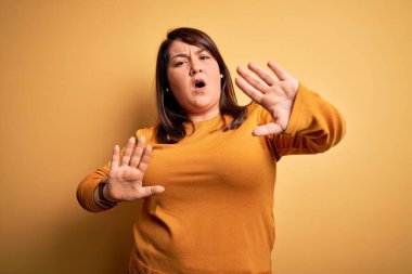 Güzel esmer ve cüsseli bir kadın izole edilmiş sarı arka plan üzerine günlük süveter giyip avuç içi hareketlerini durduruyor, kızgın ve hayal kırıklığına uğramış bir ifade takınıyor.