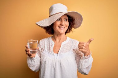 Orta yaşlı kıvırcık kadın tatilde izole edilmiş sarı arka planda bir bardak su içiyor ve gülümseyen bir gülümsemeyle baş parmağıyla onu işaret ediyor.