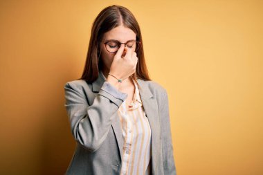 İzole edilmiş sarı arka planda ceket ve gözlük takan güzel, kızıl saçlı bir kadın yorgun burnu ve gözleri ovuşturup yorgunluk ve baş ağrısı hissediyor. Stres ve hüsran kavramı.