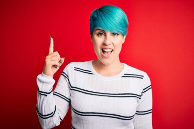 Mavi saçlı, kırmızı arka planda çizgili süveter giyen genç ve güzel bir kadın başarılı bir fikri işaret ediyor. Heyecanlı ve mutlu. Bir numara..