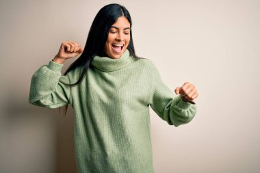 Genç ve güzel İspanyol bir kadın yeşil bir kazak giyip arka planda dans ediyor mutlu ve neşeli, hareketli ve kendinden emin bir şekilde müzik dinliyor.