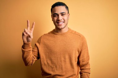 İzole edilmiş sarı arka planda dikilen sıradan süveter giyen genç Brezilyalı adam kameraya bakıp zafer işareti yapıyor. İki numara..