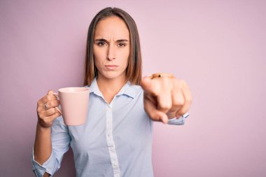 Genç ve güzel bir kadın izole edilmiş pembe arka planda durup kahve içerek kamerayı işaret ediyor ve sen, el işareti, ön taraftan pozitif ve kendinden emin bir hareket.