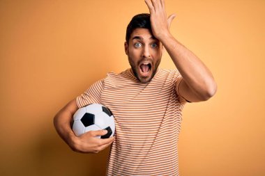 Handsome player man with beard playing soccer holding footballl ball over yellow background surprised with hand on head for mistake, remember error. Forgot, bad memory concept. clipart