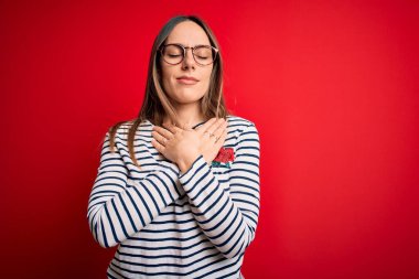 Kırmızı arka planda duran gözlüklü, mavi gözlü, elleri göğsünde, gözleri kapalı ve yüzünde minnettarlıkla gülümseyen genç, güzel sarışın kadın. Sağlık kavramı.