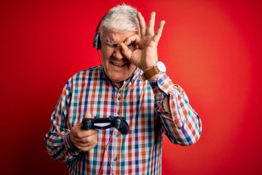 Kıdemli yakışıklı, ağarmış oyuncu oyun oynuyor, joystick ve kulaklık kullanıyor, gülümsüyor, el pençe divan durup parmaklarıyla işaret veriyor.