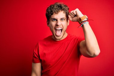 Sarışın, kıvırcık saçlı, kırmızı arka plan üzerine gündelik tişört giyen sinirli ve öfkeli genç bir adam öfkeyle bağırırken yumruğunu kaldırıyor. Öfke ve agresif kavram.