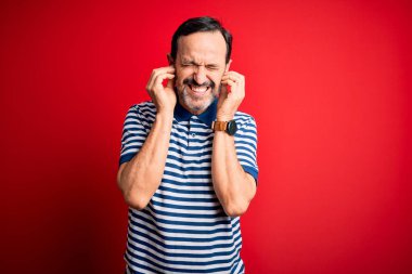 Orta yaşlı, beyaz, sıradan çizgili polo giyen, izole edilmiş kırmızı arka planda duran, yüksek sesli müzik sesi için rahatsız edici ifadeleri olan kulakları parmaklarıyla kapatan bir adam. Sağır kavramı.