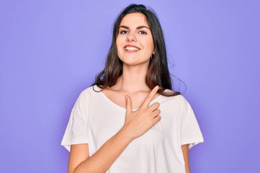 Genç, güzel, esmer bir kadın mor arka plan üzerine günlük beyaz tişört giyiyor. Gülümsüyor ve mutlu ve doğal bir ifadeyle elini ve parmağını kaldırıyor.
