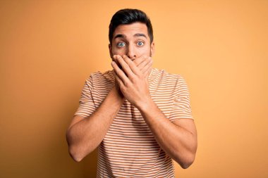Sarı arka plan üzerine sıradan çizgili tişört giyen sakallı yakışıklı genç adam ağzını yanlışlıkla ellerle kapatarak şok etti. Gizli konsept.