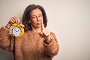 Orta yaşlı esmer kadın izole edilmiş arka planda klasik alarmlı saati tutuyor. Parmağını kaldırıyor ve kızgın bir ifade sergiliyor.