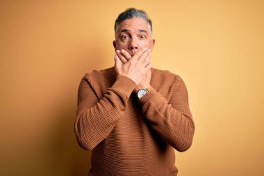 Sarı arka plan üzerine sıradan süveter giyen orta yaşlı, yakışıklı, gri saçlı bir adam ağzını yanlışlıkla ellerle kapatarak şok etti. Gizli konsept.