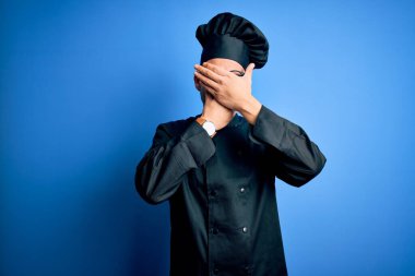 Genç, yakışıklı aşçı üniforması ve izole edilmiş mavi arka planda şapka giyen gözleri ve ağzı kapalı, şaşırmış ve şok olmuş bir şekilde. Duygularını gizliyor.