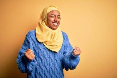 Genç Afrikalı Afro kadın, izole edilmiş sarı arka plan üzerine Müslüman başörtüsü takıyor çok mutlu ve heyecanlı bir şekilde kollarını kaldırıp, gülümseyerek ve başarı için çığlık atarak kazanan jestler yapıyor. Kutlama konsepti.