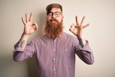 İzole edilmiş arka planda gözlük takan, sakallı, yakışıklı, kızıl saçlı yakışıklı iş adamı. Gözleri kapalı, meditasyon yaparken parmaklarıyla gülümsüyor. Yoga konsepti.