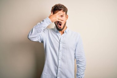 Sakallı, çizgili tişörtlü genç bir adam beyaz arka planda dikiliyor. Şok olmuş bir şekilde yüzünü ve gözlerini kapıyor. Utanmış bir ifadeyle parmaklarının arasından bakıyor..