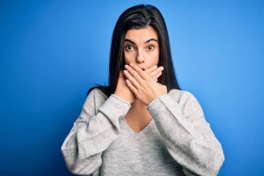 Young beautiful brunette woman wearing casual sweater standing over blue background shocked covering mouth with hands for mistake. Secret concept.