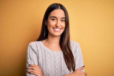 Sarı arka planda beyaz süveter giyen, kollarını kavuşturup kameraya gülümseyen güzel esmer kadın. Pozitif kişi.