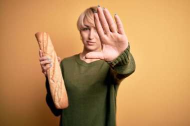 Sarışın, kısa saçlı genç bir kadın sarı arka planda taze ekmek ekmeği tutuyor. Eli açık, dur işareti yapıyor. Ciddi ve kendinden emin bir ifadeyle, savunma hareketi yapıyor.