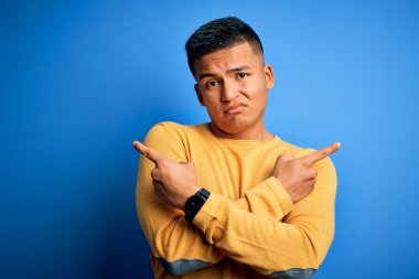 İzole edilmiş mavi arka plan üzerinde sarı süveter giyen genç, yakışıklı latin adam iki tarafı da işaret ediyor, farklı yönler farklı.