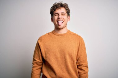 Young blond handsome man with curly hair wearing casual sweater over white background sticking tongue out happy with funny expression. Emotion concept.