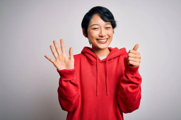 年轻美丽的亚洲女孩穿着宽松的运动衫 头戴白色背景的帽衫 头戴六号手指 面带微笑 自信而快乐 — 图库照片