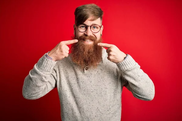 英俊的爱尔兰红头发男人 留着胡子 身穿休闲衫 眼镜罩在红色背景上 笑容满面 用手指 牙齿和嘴指尖 牙齿健康概念 — 图库照片