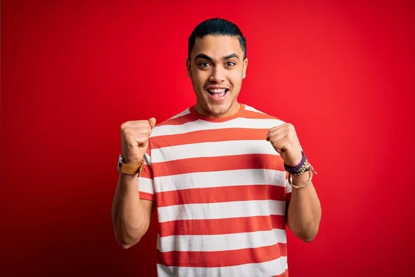 年轻的巴西男子穿着宽松的条纹T恤 站在孤立的红色背景上 以张开双臂和睁开眼睛庆祝对成功的惊奇和惊奇 赢的概念 — 图库照片
