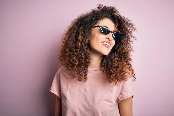 Young beautiful woman with curly hair and piercing wearing funny thug life sunglasses looking away to side with smile on face, natural expression. Laughing confident.