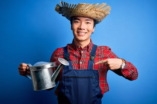 Young handsome chinese farmer man wearing apron and straw hat holding watering can with surprise face pointing finger to himself