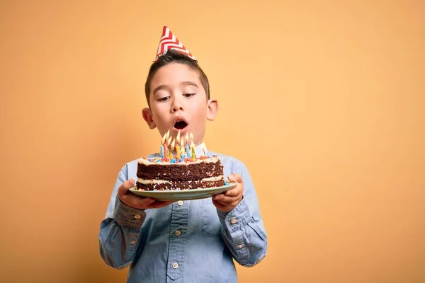 Adorable toddler wearing birthday cap holding cake with candles standing over isolated yellow background