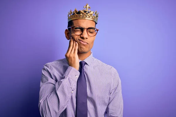 歯の痛みや歯の病気のために痛みを伴う表現で手で口に触れる紫色の背景に王の黄金の冠を身に着けている若いハンサムなアフリカ系アメリカ人男性 歯医者 — ストック写真