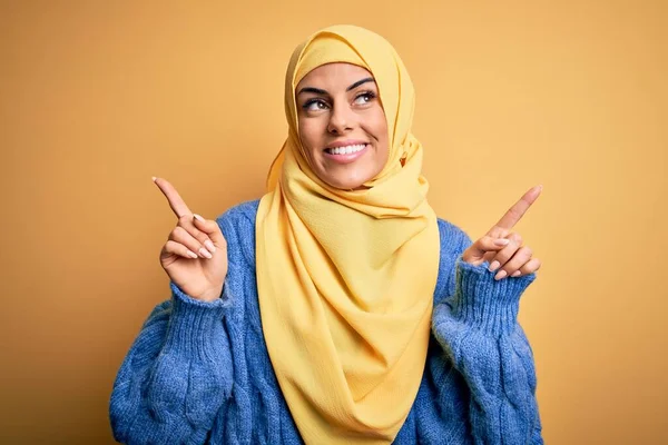 若い美しいブルネットのイスラム教徒の女性は 別の方向に指で自信を持って指して笑顔孤立した黄色の背景の上にアラブヒジャーブを身に着けています 広告のコピースペース — ストック写真
