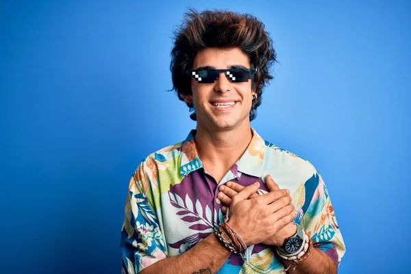 Young handsome man wearing thug life glasses standing over isolated blue background smiling with hands on chest with closed eyes and grateful gesture on face. Health concept.