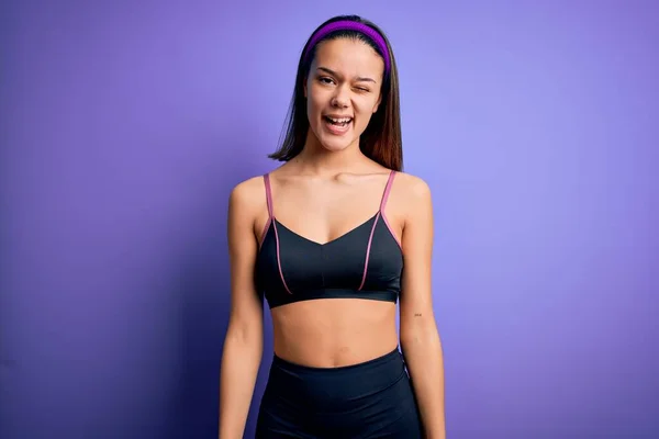 年轻而漂亮的女运动员穿着运动服 站在孤立的紫色背景上 眨着眼睛 带着性感的表情 开朗而快乐地看着摄像机 — 图库照片