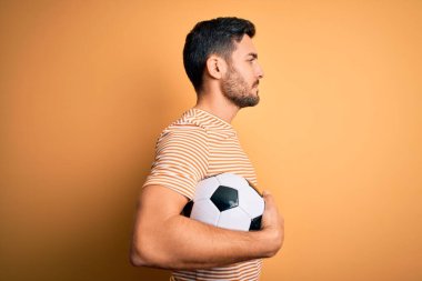 Sakallı yakışıklı oyuncu futbol topunu sarı arka planda tutarak oynuyor. Yan tarafa bakıyor, rahat profil pozu veriyor ve kendine güvenen bir gülümseme takınıyor..