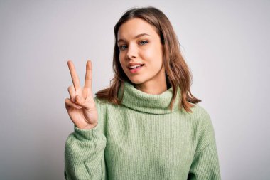 Kışlık süveter giyen genç ve güzel bir kız izole arka planda dikiliyor. Kameraya gülümsüyor ve zafer işareti yapıyor. İki numara..