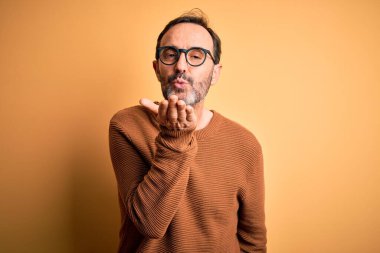 Middle age hoary man wearing brown sweater and glasses over isolated yellow background looking at the camera blowing a kiss with hand on air being lovely and sexy. Love expression. clipart