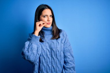 Mavi gözlü genç esmer kadın balıkçı yaka kazak giyiyor ağzı ve dudakları parmakları fermuar gibi kapalı. Gizli ve sessiz, tabu konuşması