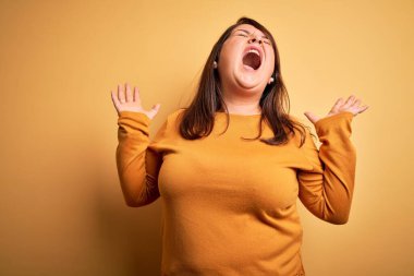 Güzel esmer ve cüsseli bir kadın izole edilmiş sarı arka plan üzerine sıradan bir kazak giyiyor çılgın ve sinirli bir şekilde bağırıyor ve agresif bir ifade ve kollarla bağırıyor. Hayal kırıklığı konsepti.