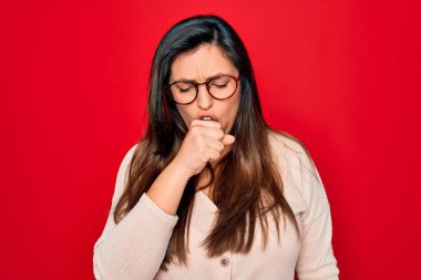 Kırmızı izole edilmiş arka planda duran gözlüklü genç İspanyol kadın kendini kötü hissediyor ve soğuk algınlığı veya bronşitin belirtisi olarak öksürüyor. Sağlık hizmeti kavramı.