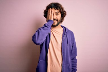 Genç, yakışıklı, sakallı, pembe arka planda rahat bir kazak giyen, bir gözünü elinle kapatan, yüzünde kendine güvenen bir gülümseme ve sürpriz duygular besleyen yakışıklı bir adam..