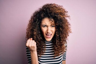 Kıvırcık saçlı, piercingli, normal çizgili tişört giyen kızgın, kızgın ve öfkeli genç bir kadın öfkeyle bağırırken yumruğunu kaldırıyor. Öfke ve agresif kavram.