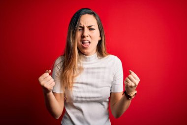 Güzel sarışın kadın, mavi gözlü, kırmızı arka plan üzerine gündelik beyaz tişört giymiş kızgın ve kızgın yumruklarını kaldırırken öfkeyle bağırıyor. Öfke ve agresif kavram.