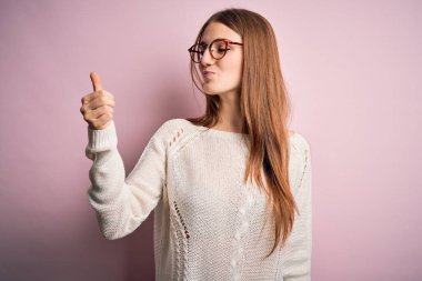 Genç, güzel, kızıl saçlı bir kadın pembe arka planda gözlük ve süveter takıyor. Gururlu görünüyor, gülümsüyor, baş parmak işareti yapıyor.