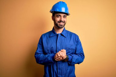 Sakallı tamirci mavi üniformalı ve sarı arka planda emniyet kaskı takmış elleri çapraz parmaklı rahat ve neşeli bir şekilde gülümsüyor. Başarı ve iyimser
