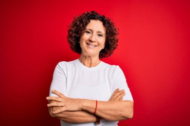 Orta yaşlı güzel kıvırcık saçlı kadın izole edilmiş kırmızı arka planda tişört giyip kollarını kavuşturup kameraya bakarken gülümsüyor. Pozitif kişi.
